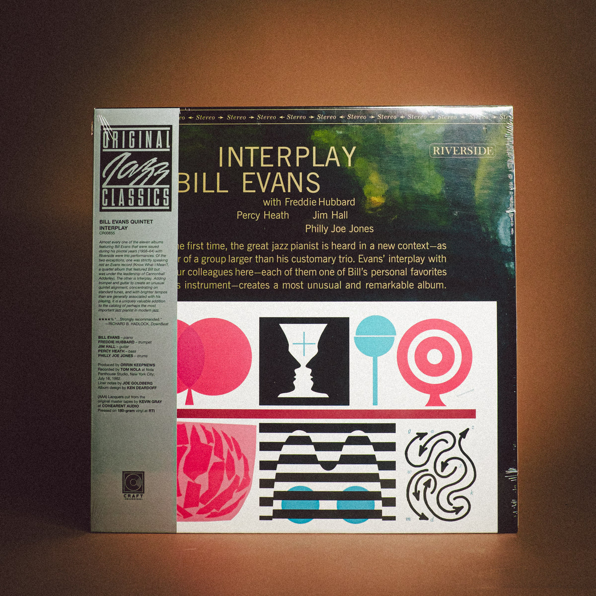 A vinyl record album cover for 'Interplay' by Bill Evans, photographed standing upright against a dark brown background. The cover features a green and black abstract design on the right and a light gray text panel on the left with track listings, plus colorful geometric graphics at the bottom.