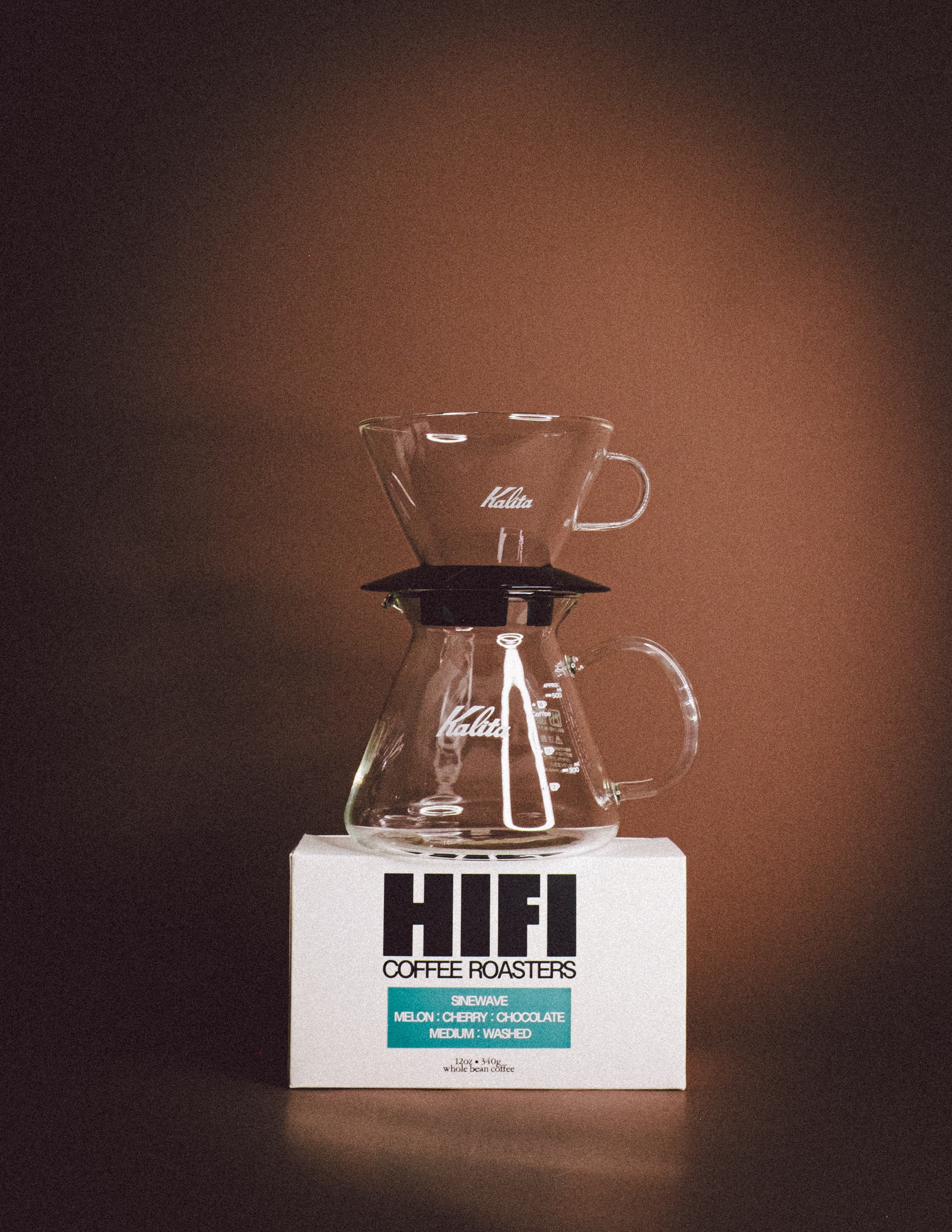 Glass coffee maker on a white block with 'HIFI Coffee Roasters' branding against a brown background