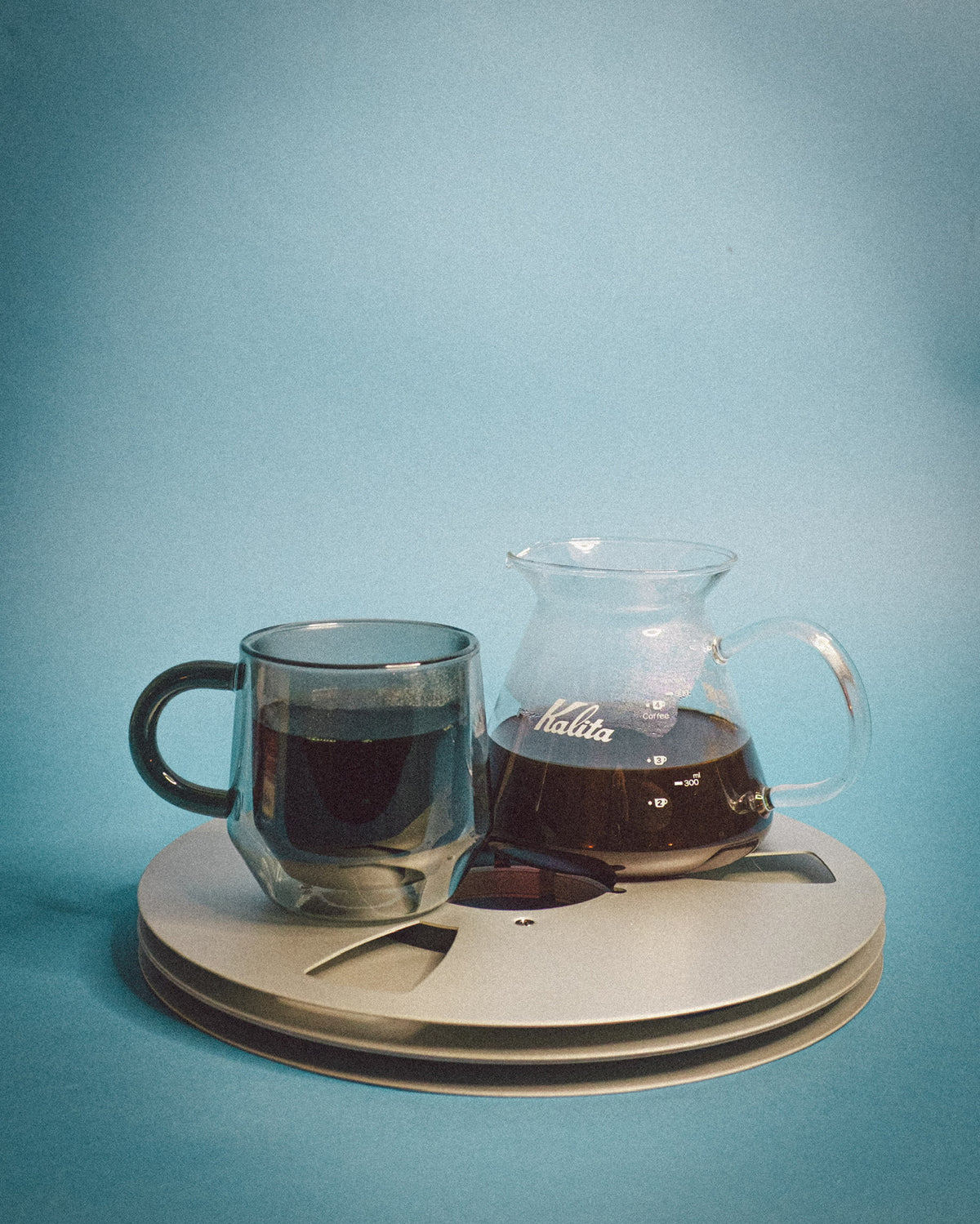 Coffee cup and Kalita coffee maker on a circular wooden coaster with a blue background
